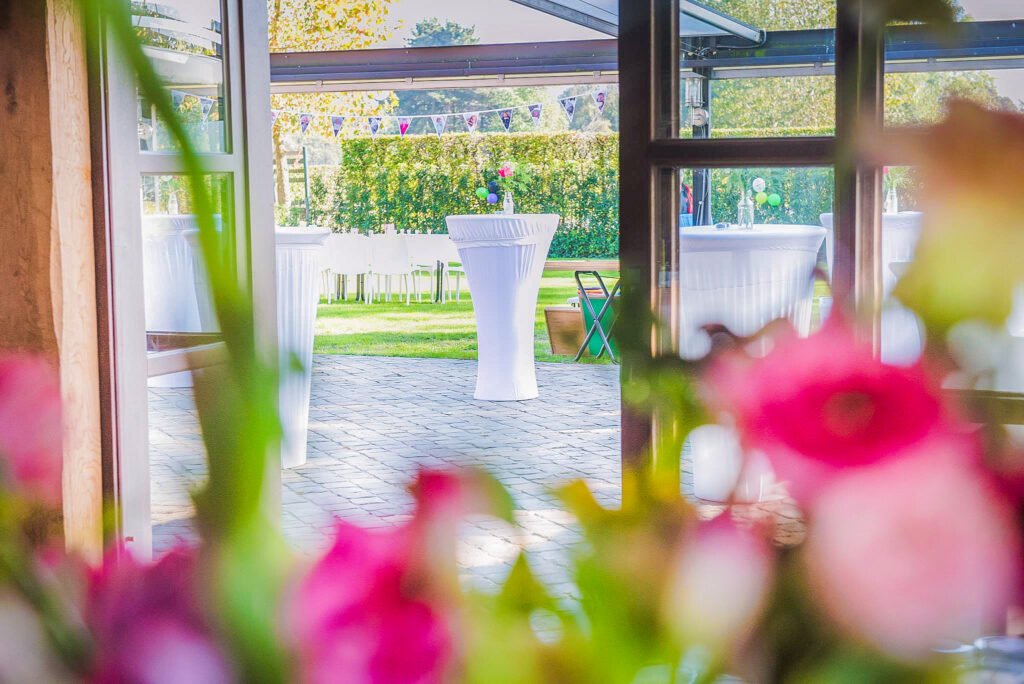 Uitzicht vanuit het gebouw op het buitenterras met witte statafels en groene tuin bij Feestlocatie Alegria