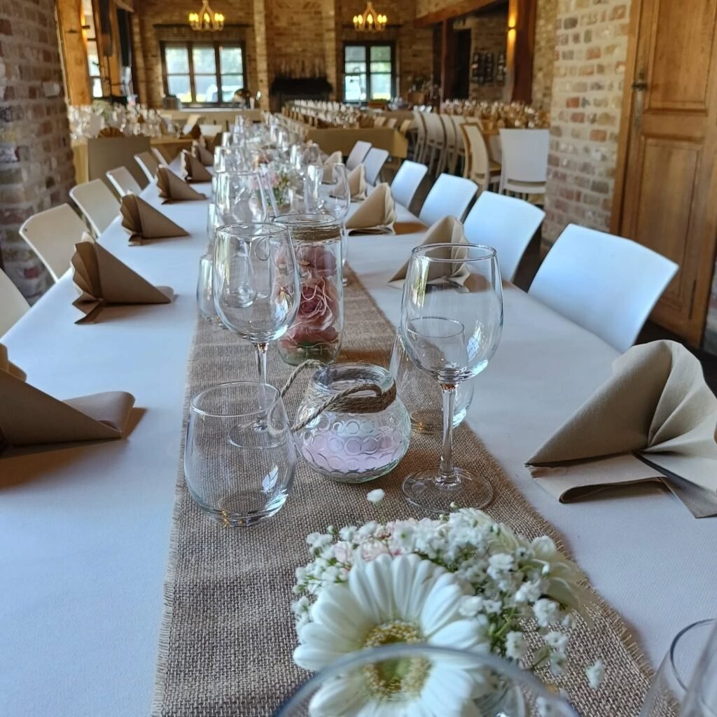 Elegant gedekte feesttafel met glazen, jute tafelloper en witte stoelhoezen in de rustieke zaal van Feestlocatie Alegria